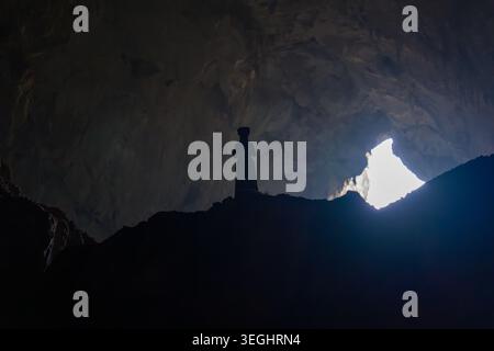 Silhouette stands inside vast cave. Light floods in from distant opening. Cave walls rise steep and dark. Figure appears small against cavern. Natural Stock Photo