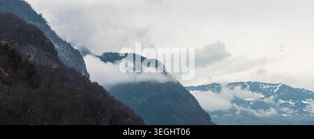 Misty Alps mountains rise above forested slopes as low clouds drift through valleys, creating a moody, serene landscape ideal for travel, nature, and Stock Photo