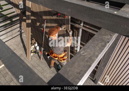 A worker is busy installing wooden panels on a structure at a construction site. The worker stands on a roof, using tools and adjusting the panels car Stock Photo