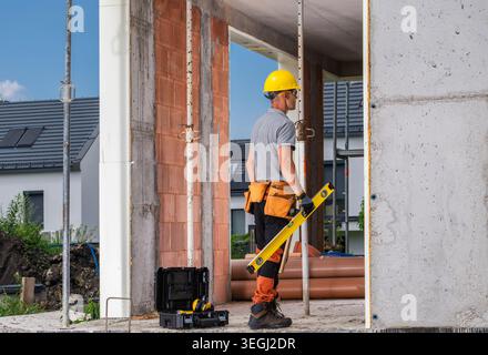 A worker stands at a construction site holding a level tool in one hand. He wears a yellow hard hat and work clothing. Tools are set nearby. The sun i Stock Photo