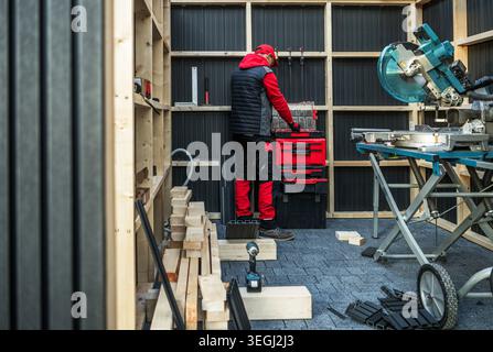 A man stands in a small workshop. He organizes red toolboxes and adjusts his equipment. The space has wooden walls and various tools on display. Light Stock Photo