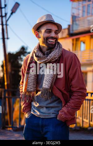 A smiling man with a beard, wearing a hat and scarf, stands outdoors with buildings in the background in Chakrata, Uttarakhand, India. Stock Photo