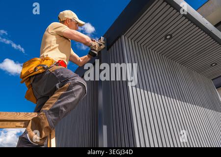 A worker is standing on a ladder while installing panels on a building. The worker is focused on the task. It is a sunny day with few clouds in the sk Stock Photo