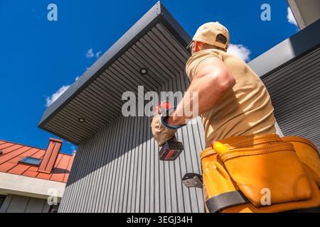 A worker stands with a tool in hand, focused on installing lighting on the exterior of a building. The worker wears a tan shirt and a tool belt while Stock Photo