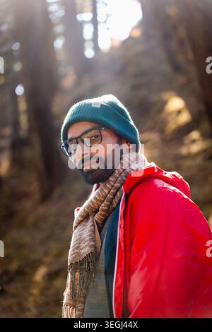 A bearded man wearing a green beanie, glasses, and a red jacket stands in a sunlit forest in Chakrata, Uttarakhand, India. Stock Photo