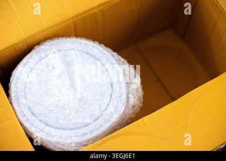 Close up view of round object securely wrapped in protective bubble wrap resting inside large brown cardboard shipping box ready for safe transport an Stock Photo
