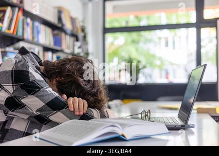 Exhausted teenage student in a plaid shirt sleeping on books at a library desk with an open laptop. Tired male teenager passed out from studying Stock Photo