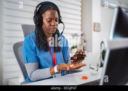Nurse wearing headset pouring pills while working on computer Stock Photo
