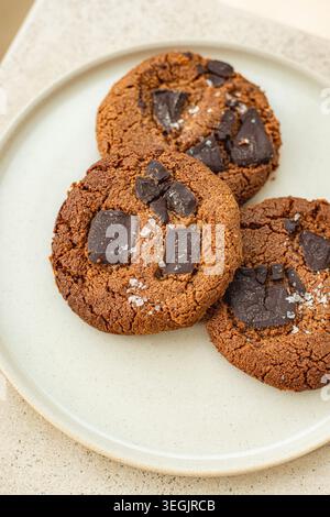 A detailed macro shot of three freshly baked chocolate chunk cookies sprinkled with coarse sea salt flakes, stacked on a white ceramic plate. Stock Photo