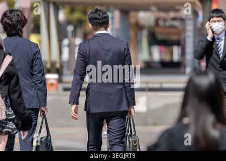 Sales reps in dark suits carrying business bags walking along bustling plaza as other people pass nearby, reflecting the daily corporate rhythm. Stock Photo