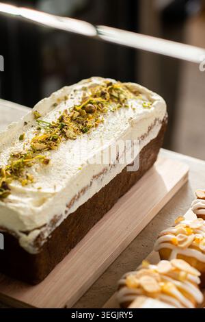 A close-up shot of a rustic pound loaf cake covered in white cream frosting, garnished with crushed pistachio nuts and grated citrus zest on a wooden Stock Photo
