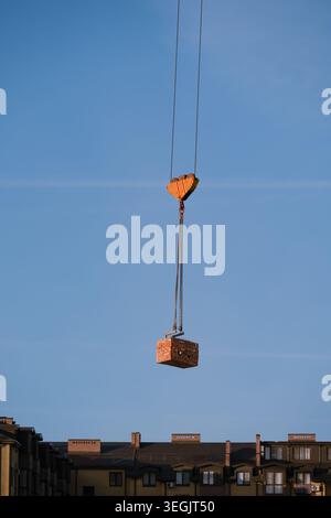 Construction crane lifting a stack of red clay bricks on steel chains against a blue sky background, with residential apartment buildings below. Stock Photo