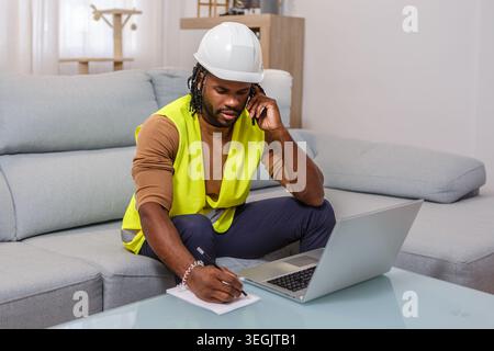 A construction worker wearing a safety vest and a hard hat sits on a couch, engaged in a phone call, taking notes with a laptop nearby in a well-lit h Stock Photo