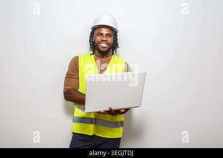 A construction worker wearing a hard hat and safety vest stands confidently, holding a laptop. He smiles broadly, showcasing a positive attitude in a Stock Photo