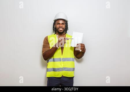 A construction worker wearing a safety vest and helmet stands indoors, pointing to a blank notepad he holds. He looks engaged and ready to present ide Stock Photo