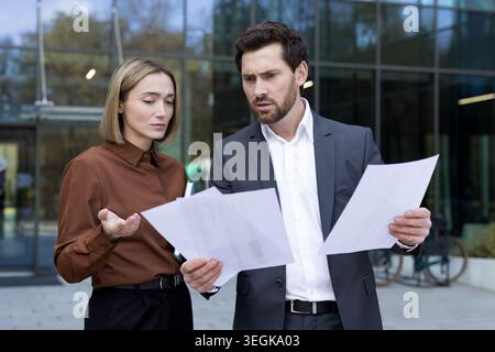 Two business professionals standing outside an office building are reviewing paperwork with frustrated expressions, discussing issues and challenges r Stock Photo