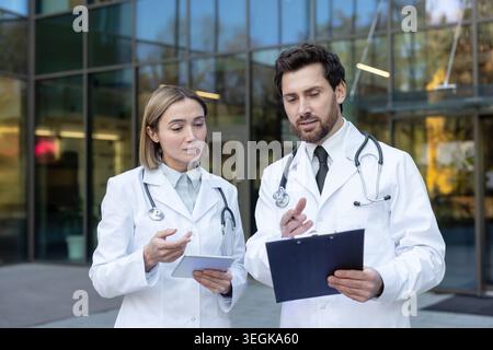 Two healthcare professionals wearing stethoscopes and lab coats discuss patient information, collaborating with a tablet and clipboard in front of a m Stock Photo