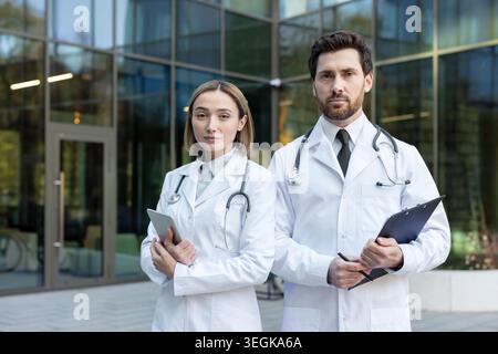Medical professionals wearing white lab coats and stethoscopes, a man holding a clipboard and pen, a woman holding a tablet, standing in front of a co Stock Photo