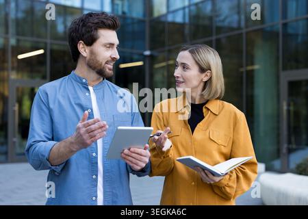 Two business professionals standing outside a modern office building, discussing project details and sharing ideas while holding a digital tablet and Stock Photo