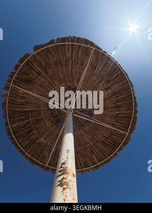 Low angle look up view of a straw beach umbrella against a clear blue sky with bright summer sun flare. Stock Photo