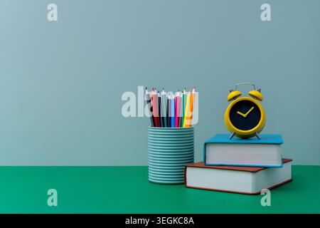 A yellow alarm clock on stacked books beside a container of colored pencils. Education, time management, academic schedules, deadlines Stock Photo