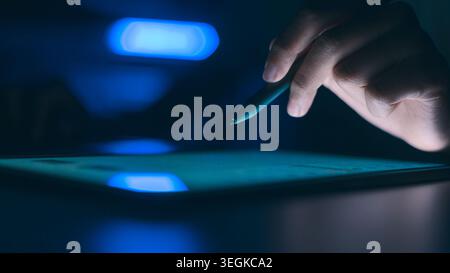 Hand holding stylus pen over digital tablet in the dark. Professional designer or artist working at night with creative blue glowing light and copy sp Stock Photo