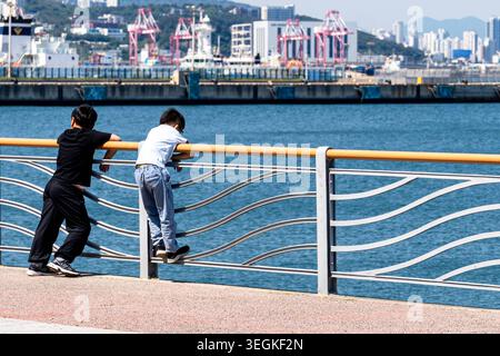 Busan, South Korea May 16, 2026: Two children are leaning against a railing on the Busan waterfront, looking out over the harbor basin. In the background are port facilities, cranes, and the maritime infrastructure of the South Korean coastal city *** Zwei Kinder lehnen an einem Geländer an der Uferpromenade von Busan und blicken auf das Hafenbecken. Im Hintergrund liegen Hafenanlagen, Kräne und die maritime Infrastruktur der südkoreanischen Küstenstadt Stock Photo