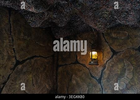 A vintage, rustic lantern hangs in a dark cave or grotto, providing warm yellow light that illuminates the surrounding textured rock walls Stock Photo