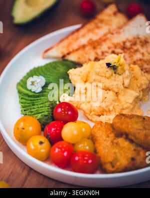A breakfast platter with scrambled eggs, turkey bacon, toast, sliced avocado, and cherry tomatoes on a wooden table Stock Photo