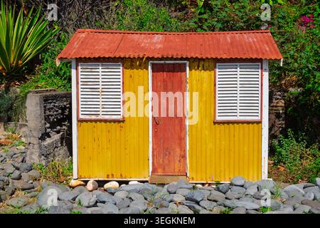 A yellow corrugated metal building with a red roof a red door and two white louvered windows stands among green foliage and grey rocks Stock Photo