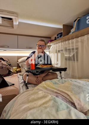 A man sitting comfortably on a bed inside a campervan, eating pizza during a camping road trip in Palmes, France. Stock Photo