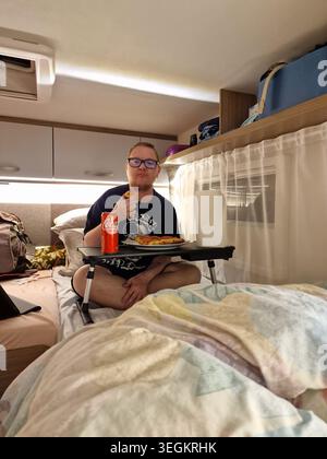A man sitting comfortably on a bed inside a campervan, eating pizza during a camping road trip in Palmes, France. Stock Photo