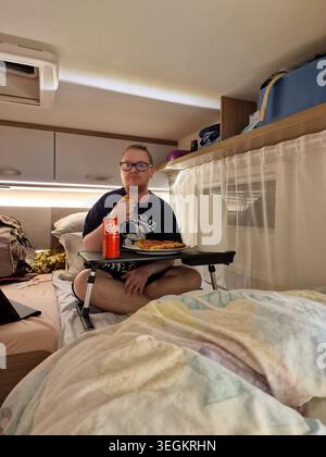A man sitting comfortably on a bed inside a campervan, eating pizza during a camping road trip in Palmes, France. Stock Photo