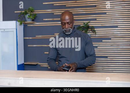Senior African American man checking smartphone, standing behind wood counter in dark zip pullover Stock Photo