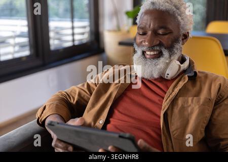 Senior African American man sitting on gray chair by window blinds, using tablet with headphones Stock Photo