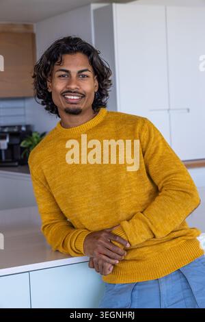 Indian man leaning on counter in kitchen, wearing mustard sweater, smiling by coffee maker Stock Photo
