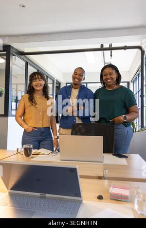 Diverse coworkers standing behind long wooden desk in modern meeting room using laptops and smiling Stock Photo
