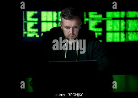 Hacker operates laptop in dark office. Hacker analyzes code screen carefully. Hacker monitors system logs for access. Hacker executes cyber attack on Stock Photo