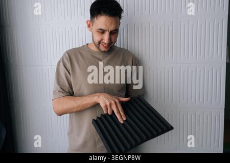 Young man holding a black acoustic foam panel, exploring soundproofing materials for studio environment, noise reduction, and audio quality Stock Photo