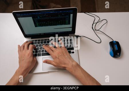 Top view of software developer typing on laptop computer with wired mouse on white desk. Managing digital audio workstation software. Illustrating Stock Photo