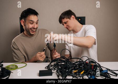 Two cheerful men engaging with challenging mess of disorganized cables and cords, finding humor in the task of identifying different connectors and Stock Photo