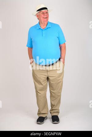 Man stands confidently in a blue polo shirt. He wears khaki trousers and a white Panama hat. His smile shows friendly warmth. Hands rest in his pocket Stock Photo