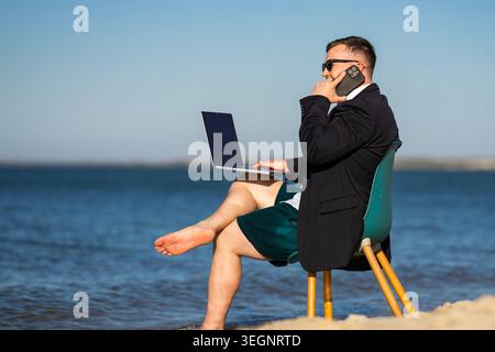Beach office shows remote work. Man works from beach chair. Beach laptop creates funny business scene. Summer work fits beach lifestyle. Stock Photo