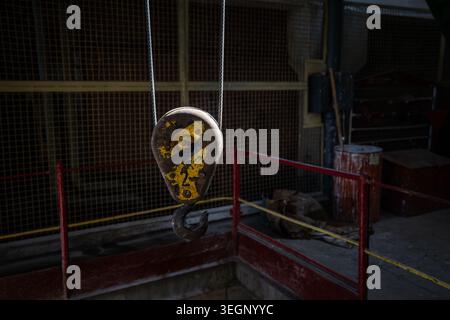 A heavily worn industrial crane hook with chipped yellow paint hangs from steel wire ropes inside a dimly lit workshop with metal railings, a mesh scr Stock Photo