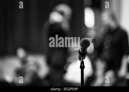 A vocal microphone stands on a mount in a dimly lit venue with blurred figures of people sitting in the background during an indoor public event or pr Stock Photo