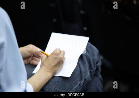 An over-the-shoulder view of a person sitting with crossed legs while writing notes on a blank sheet of white paper with a yellow wooden pencil during Stock Photo