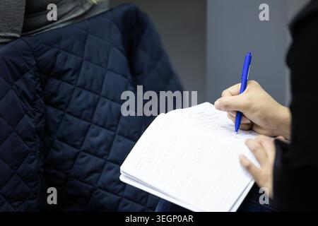 A close-up view of a person holding a blue ballpoint pen and writing notes in a lined notebook during a lecture, with another individual wearing a qui Stock Photo