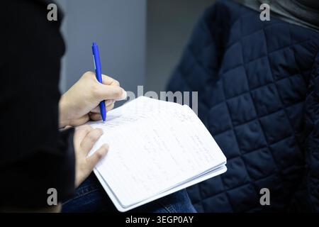 A close-up view of an individual holding a blue ballpoint pen and writing handwritten notes in a lined paper notebook while sitting indoors next to an Stock Photo