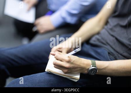 A close-up shot of a person holding a pen and a notebook while sitting during a professional seminar or business meeting with other participants seate Stock Photo