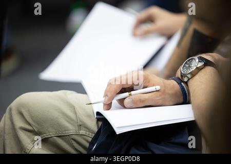 A close-up view of a person holding a white pen over an open blank notebook while sitting down during a public seminar, with another individual holdin Stock Photo